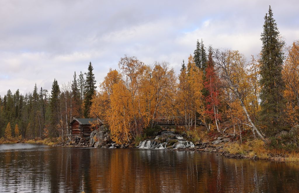 Charming autumn scene of a hut by a riverbank in Kolari, Finland with colorful trees.